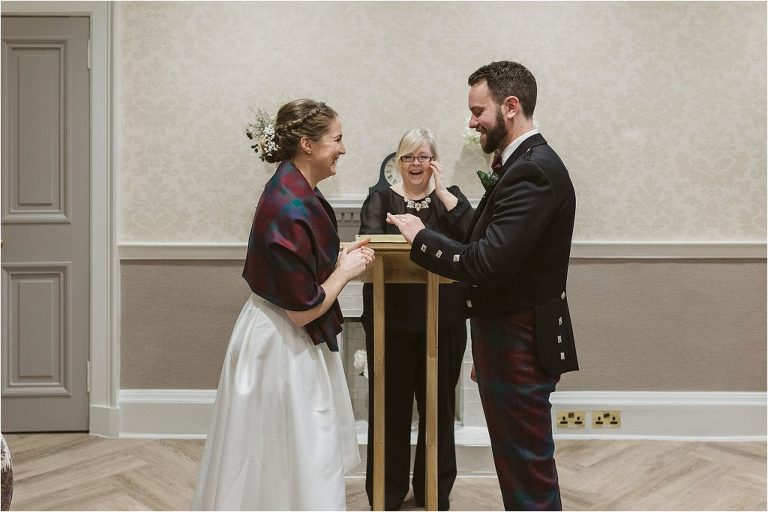 groom-inspects-wedding-ring-while-bride-is-laughing