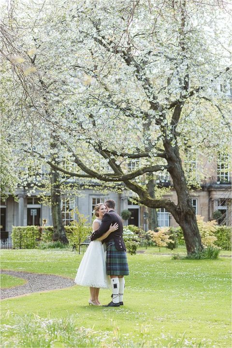 bride-and-groom-embrace-in-edinburgh-private-gardens-surrounded-by-cherry-blossom