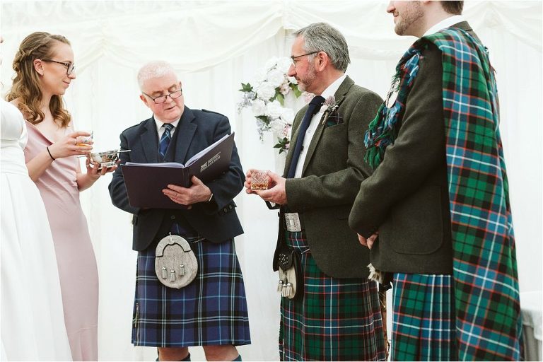 grooms-dad-and-bridesmaid-hold-glass-of-whisky-before-pouring-in-to-quaich