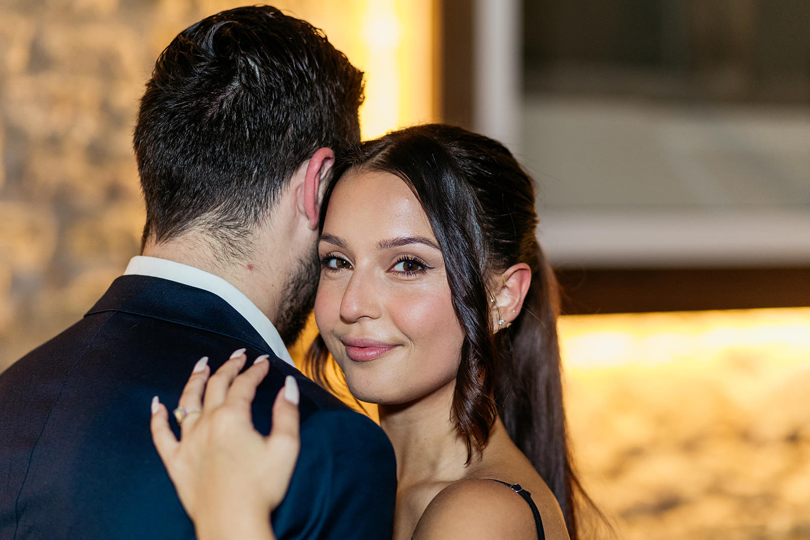 woman-looks-at-camera-and-smiles-while-her-new-fiance-hugs-her-and-leans-his-cheek-towards-her-temple