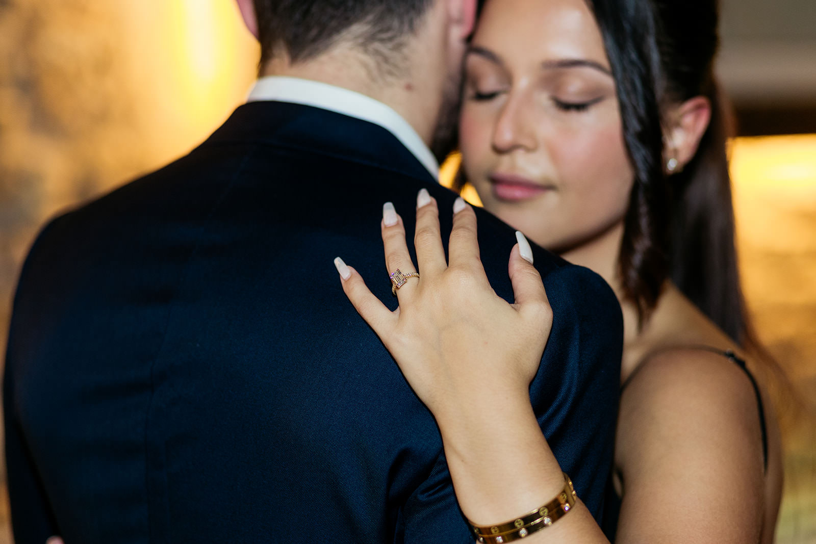 a-close-up-photo-of-womans-emerald-cut-diamond-engagement-ring-while-she-hugs-her-new-fiance-and-closes-her-eyes