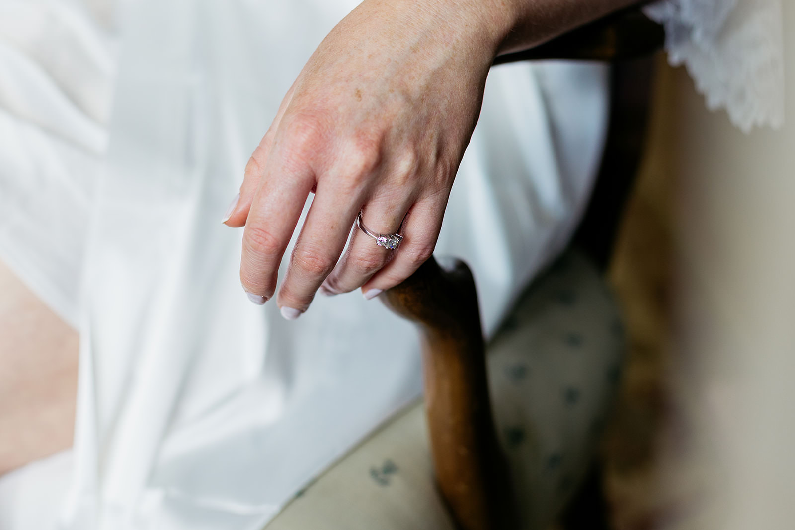 close up of a bride's hand as it rest on the wooden arm of a chair. you can see her three diamond engagement ring on her finger