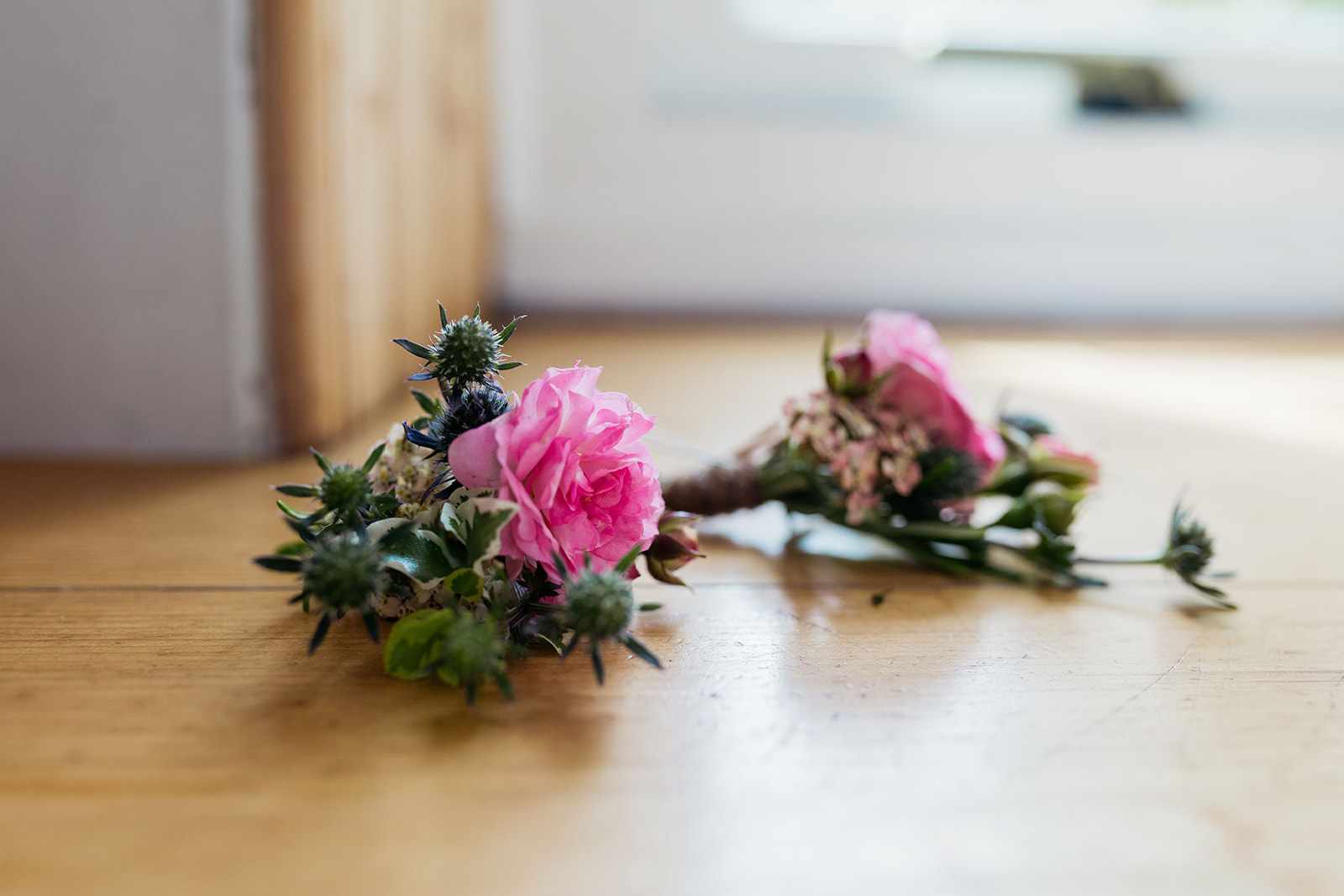 a small pink carnation and miniature thistles make up a button hole. two are lying on a wooden window sill.