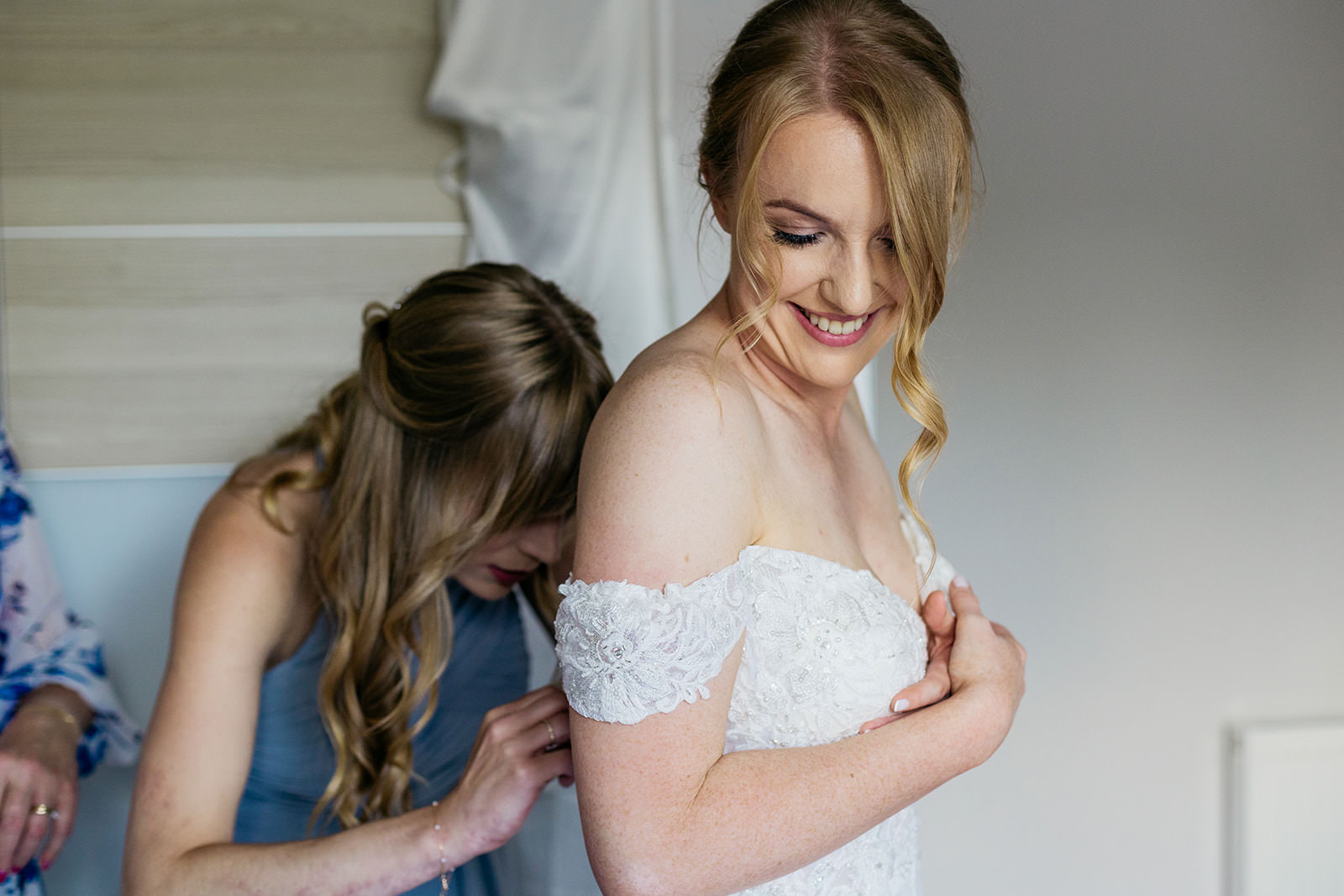 a woman is smiling as her bridesmaid helps fasten up the back of her wedding dress.