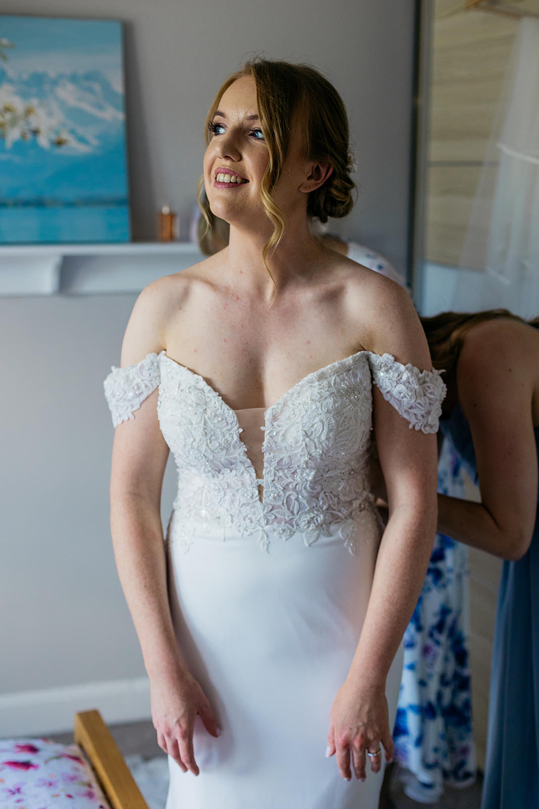 a woman is standing looking towards the window light smiling, as she gets fastened in to her dress.