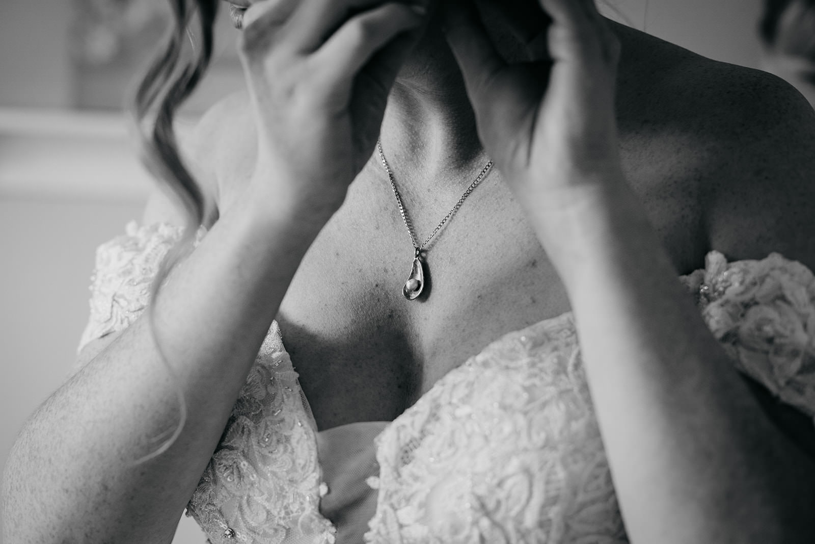 the brides necklace looks like a small muscle shell with a pearl towards the bottom. the brides hands are up towards her ear as she puts an earring on