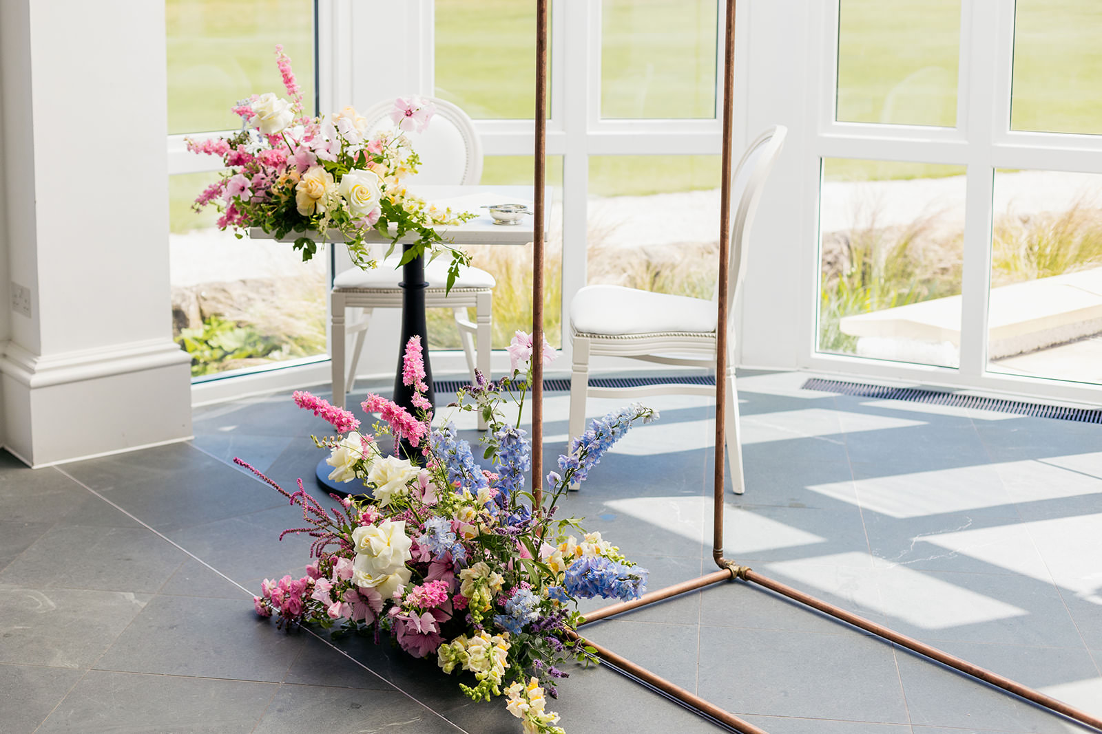 two floral arrangements, one is on the floor at the bottom of a copper frame, the other is on the corner of the singing table.