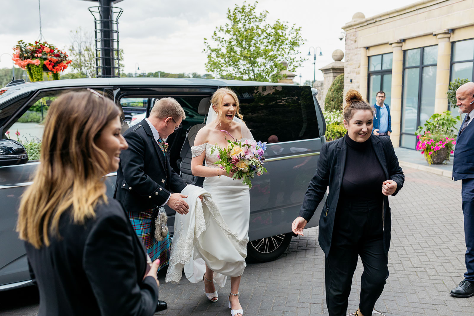 the bride and her dad arrive at the old course hotel and have gotten out of a silver people carrier. staff members have gathered to greet them.