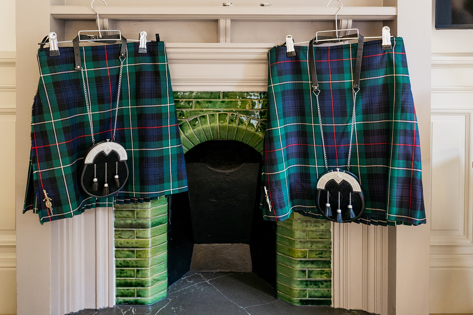 two tartan kilts with sporrans are hanging on a fireplace. there are green tiles behind them on the fireplace