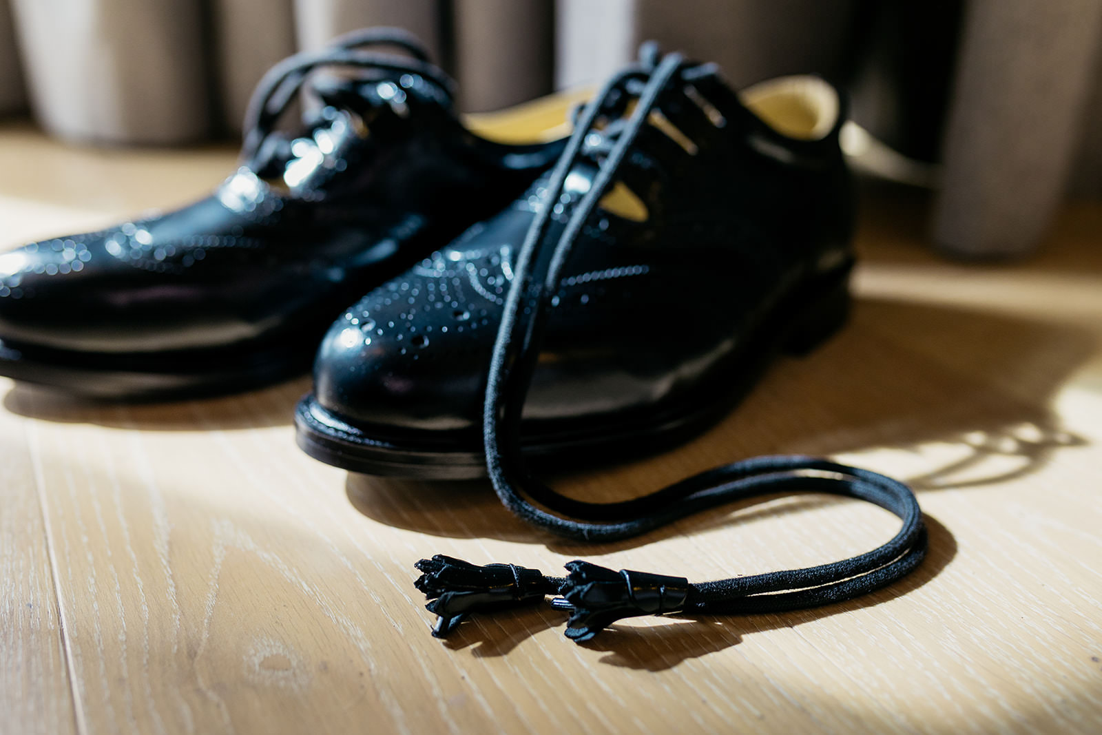 a pair of black kilt shoes are sitting on a wooden floor in the sunlight. the laces are draped in front of the shoe.