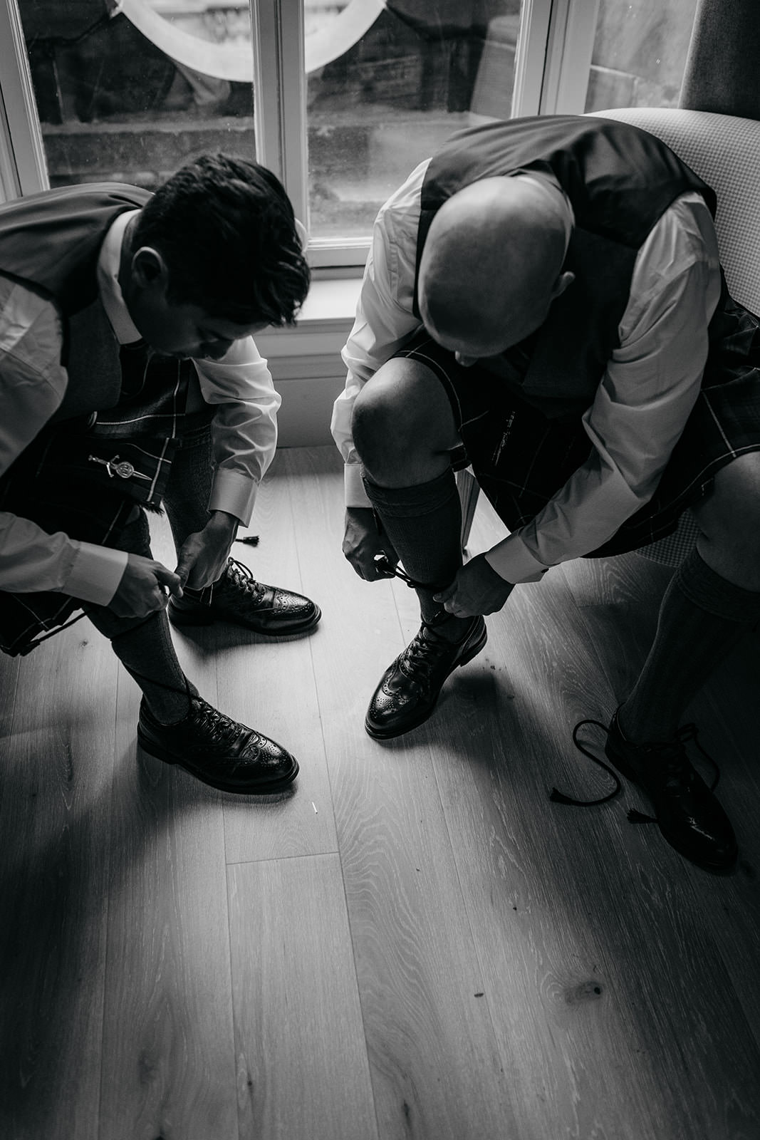 two grooms are sitting and tying up their kilt shoe laces.