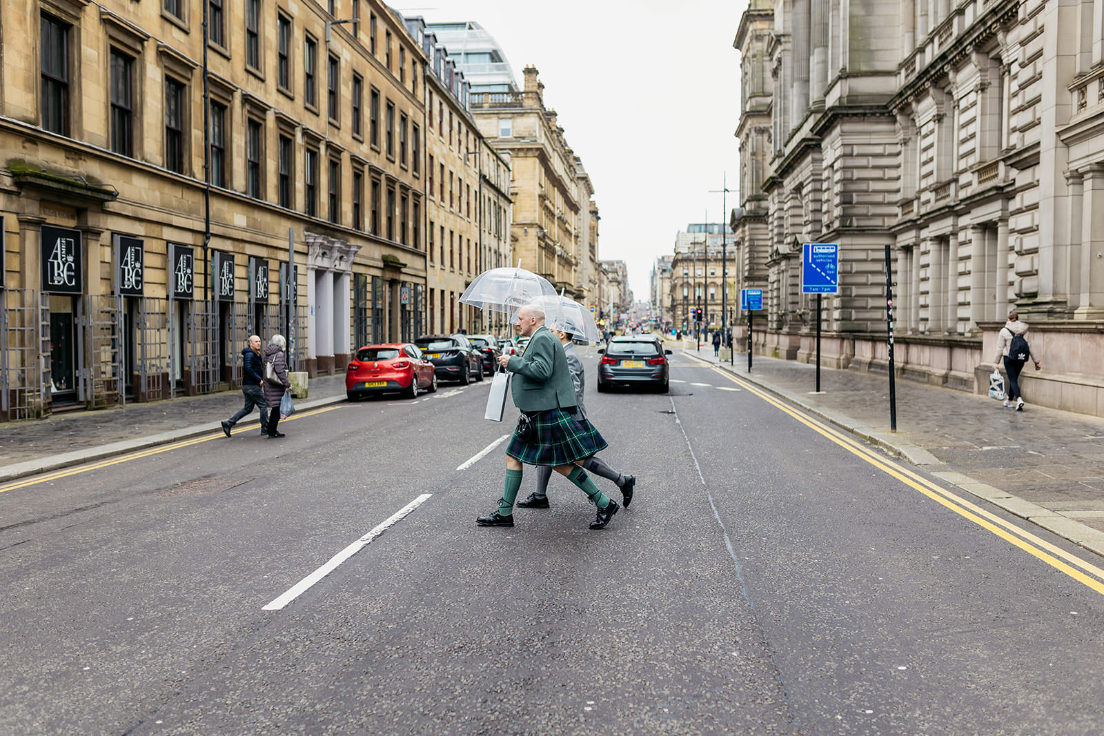 two grooms are crossing the road in glasgow city centre. they are holding clear umbrellas