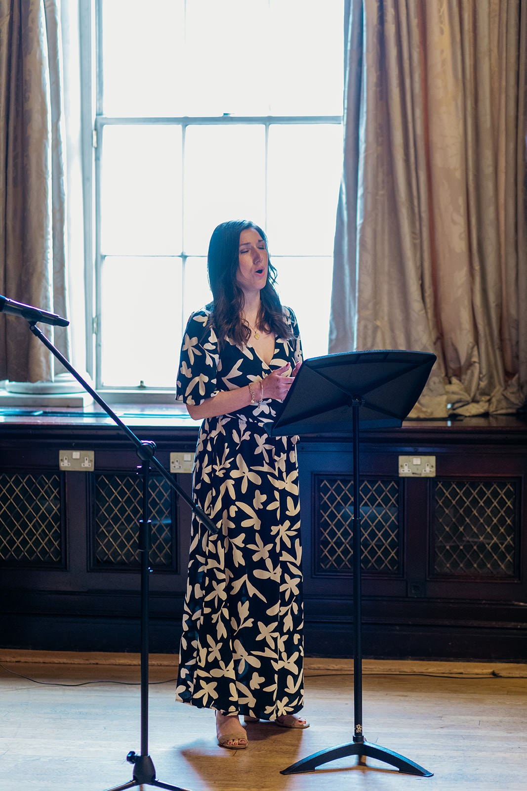 a woman wearing a black dress with a leafy design is standing and singing. there is music stand in front of her.