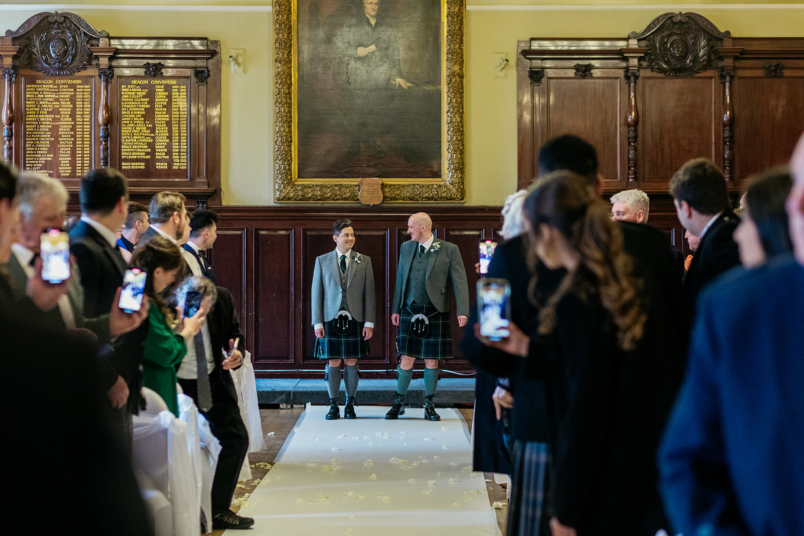 two grooms are at the bottom of the aisle at the Trades Hall in Glasgow. the wedding guests are turned to watch them.