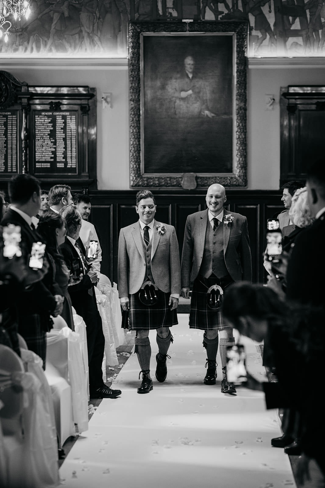 a gay couple are walking side by side down the aisle at their wedding. the guests are turned to watch them. a large oil painting portrait is hanging on the wall at the back of the room.