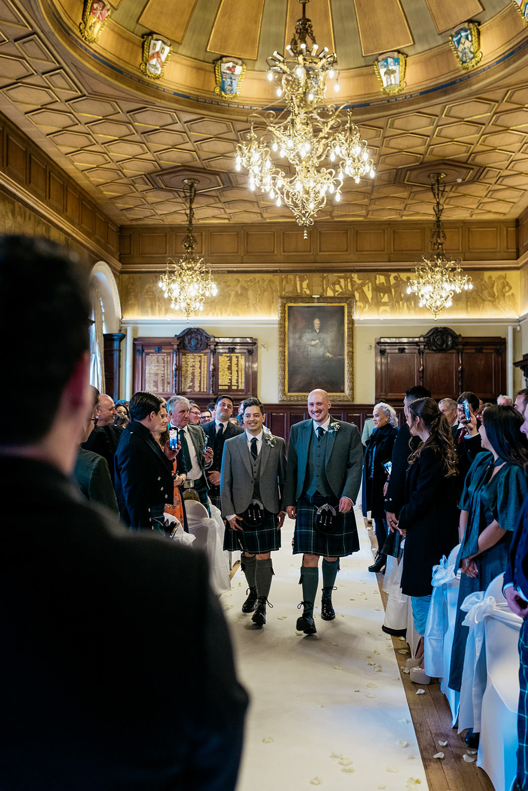 a gay couple are walking side by side down the aisle at their wedding. the guests are turned to watch them. a large oil painting portrait is hanging on the wall at the back of the room.
