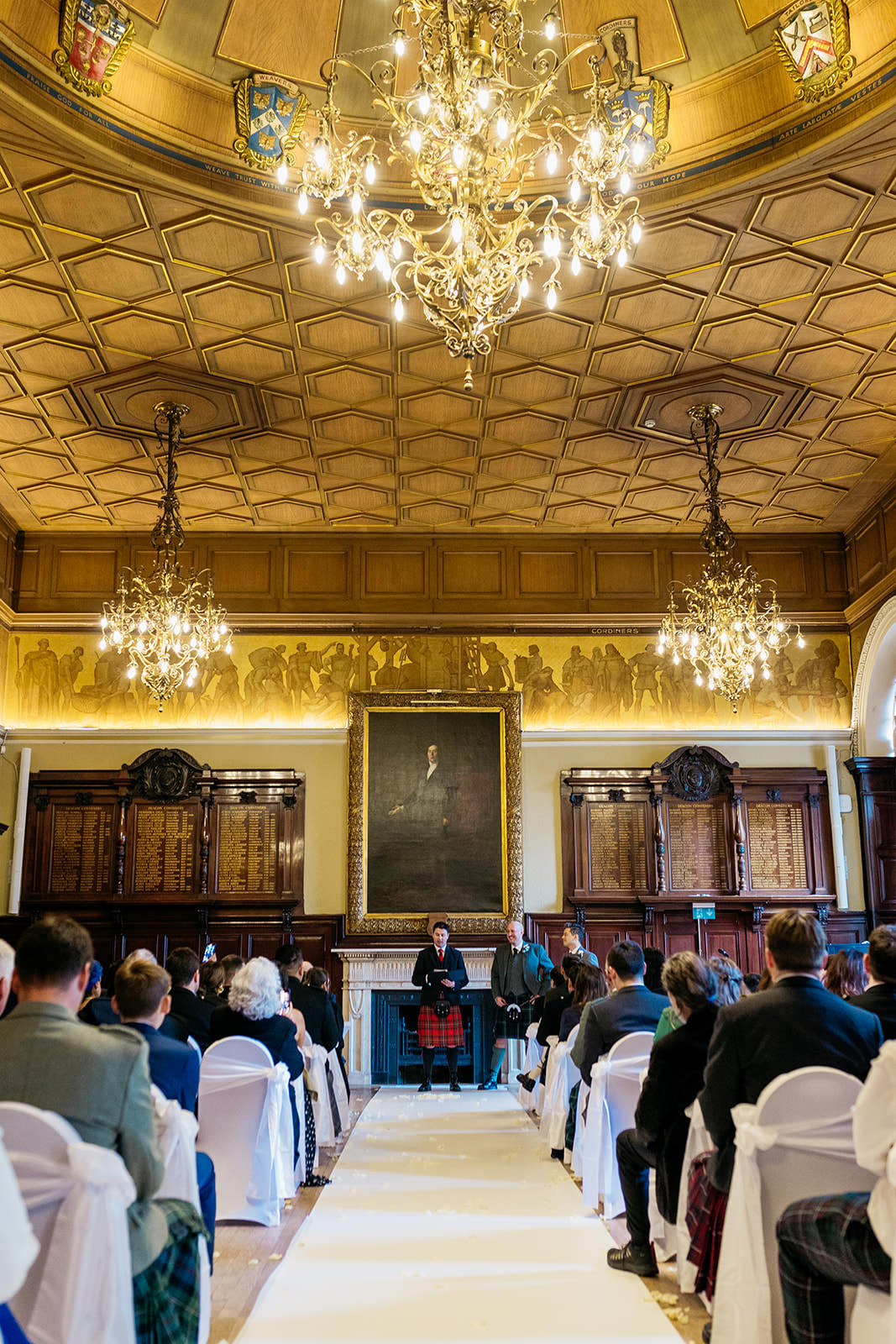 the two grooms are standing together at the top of the aisle. the guests looking at the celebrant. there are three chandeliers hanging on the gold ceiling above them.