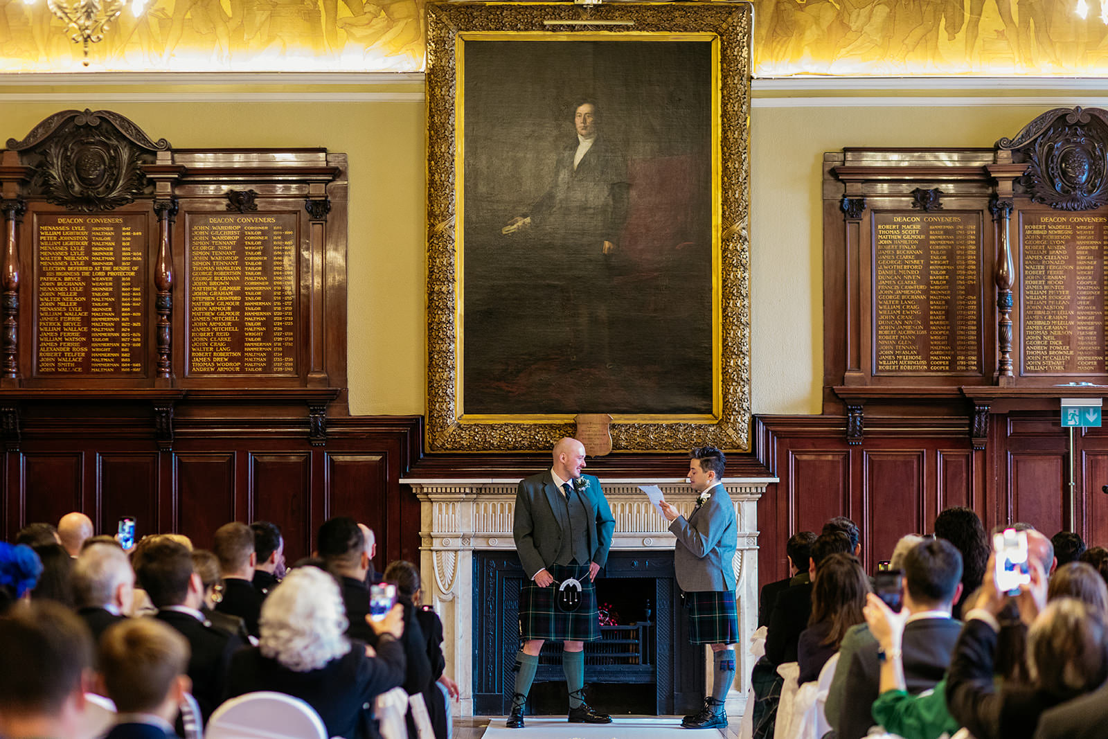 two grooms are standing and one groom is reading from a piece of paper. a large oil painting is hanging above the fireplace behind them.