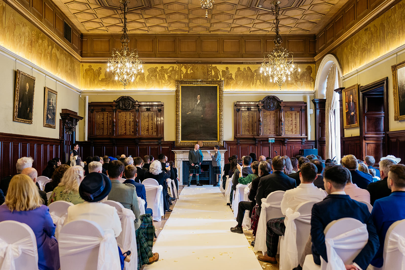 two grooms are standing at the end of their wedding aisle. one groom is reading from a piece of paper. the guests are watching them