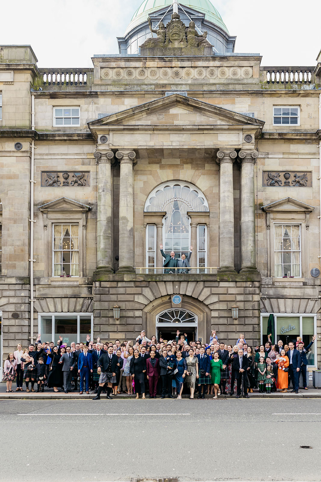 wedding guests are gathered outside of the trades hall building in glasgow. the two grooms are standing on the balcony.