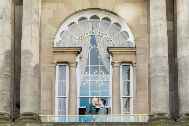 a gay couple are standing on the balcony of trades hall in glasgow kissing.one groom is wearing a green kilt jacket and the other groom is wearing a grey kilt jacket.