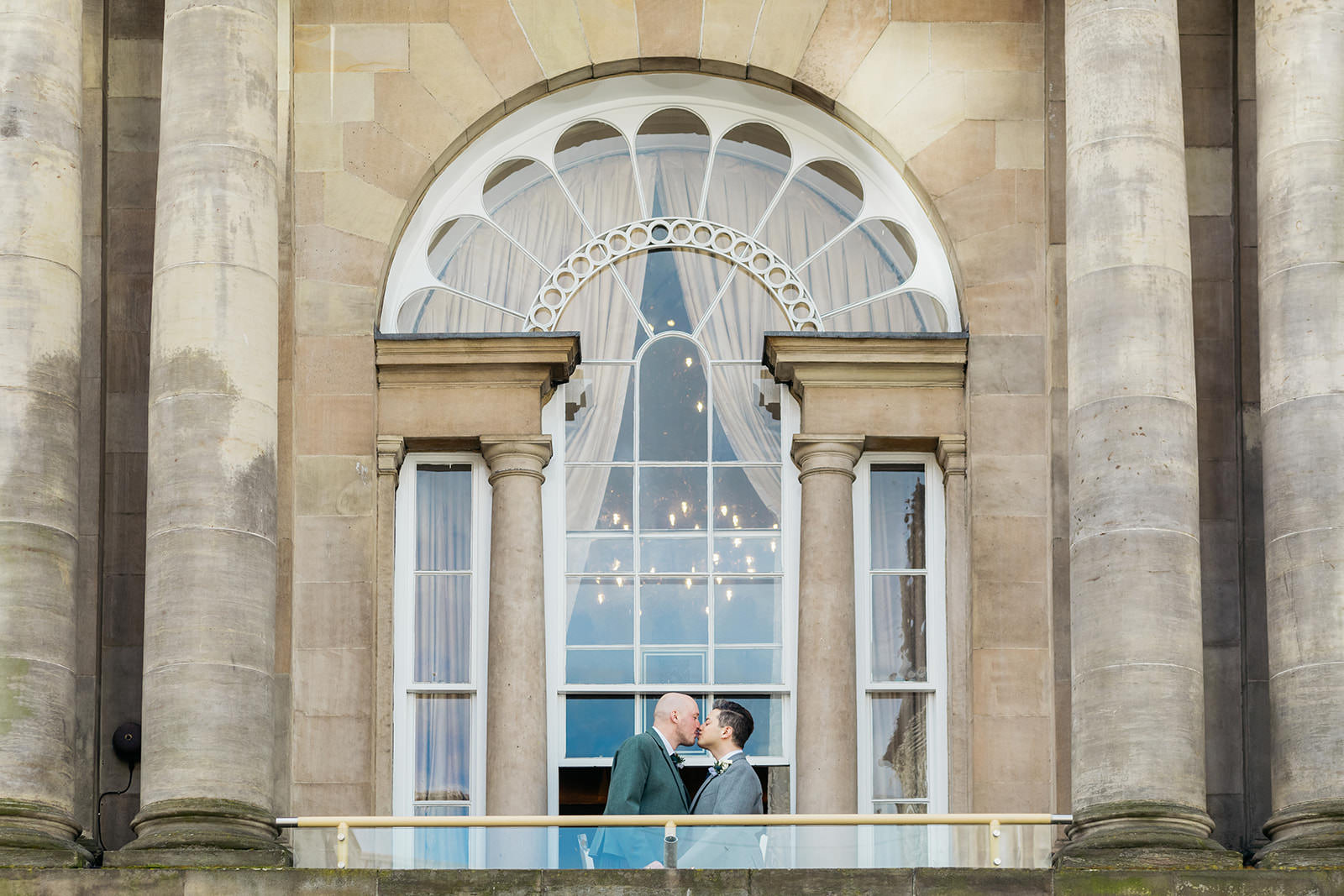 a gay couple are standing on the balcony of trades hall in glasgow kissing.one groom is wearing a green kilt jacket and the other groom is wearing a grey kilt jacket.