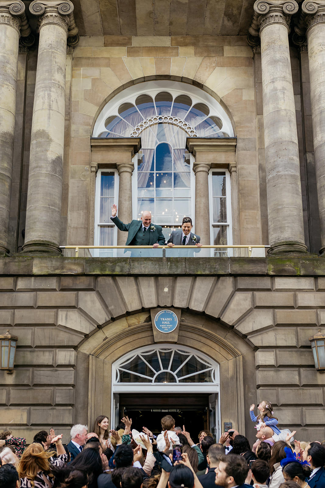 a couple are standing on the balcony of the Trades Hall. they are looking down at their guests that are gathered on the street below and waving at them.