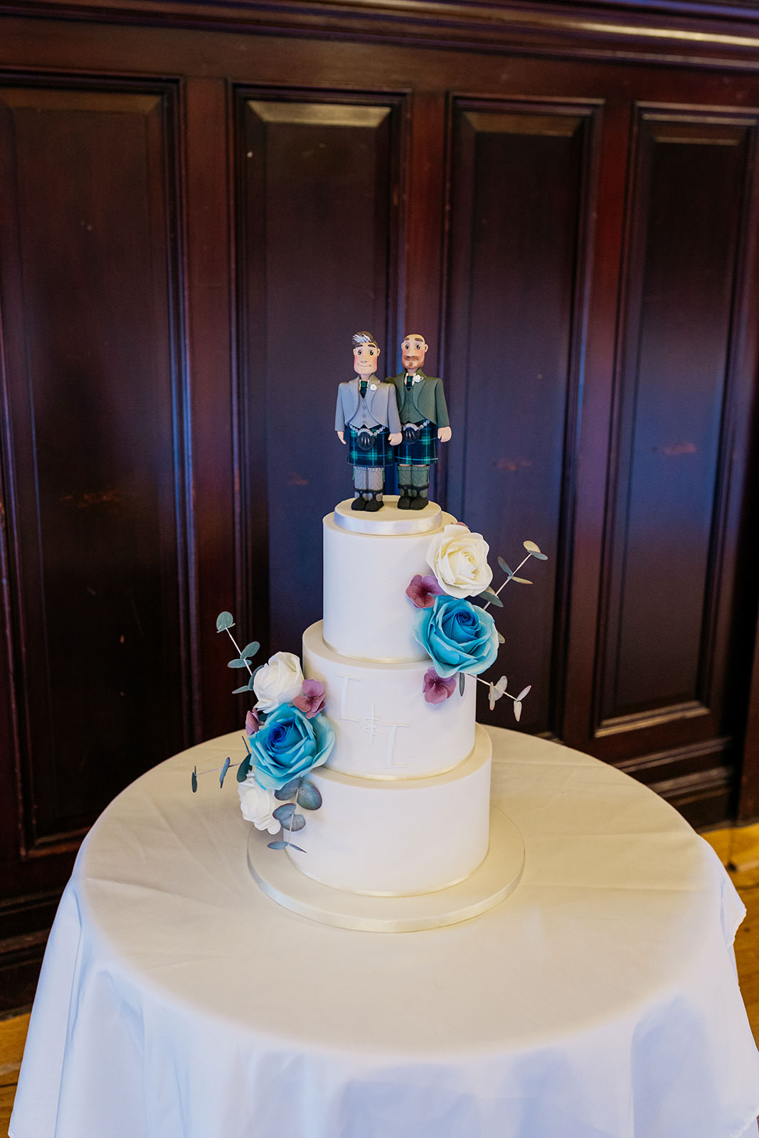 a white cake made by April Cottage Cakery is sitting on a white table cloth. it is topped with two grooms wearing kilt outfits. a blue and white flower is on each side of the three tiers.