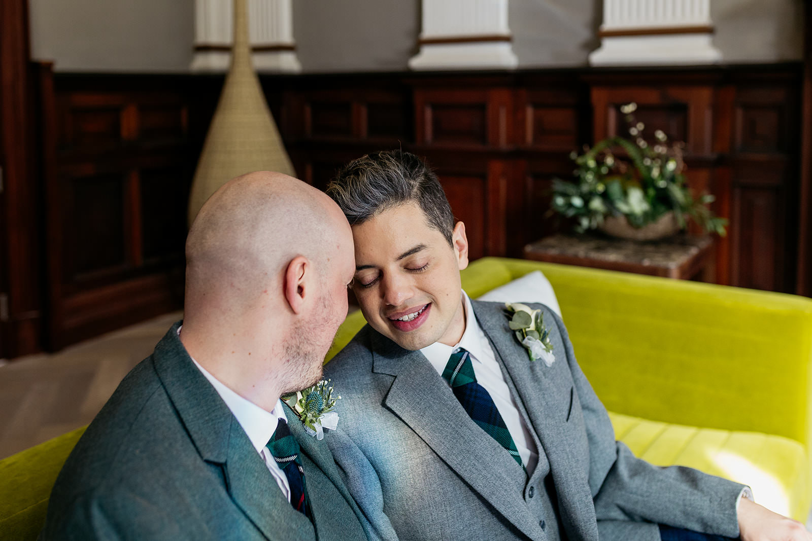 two grooms are sitting together on a green sofa. their foreheads are resting against each others and they have their eyes closed while smiling.