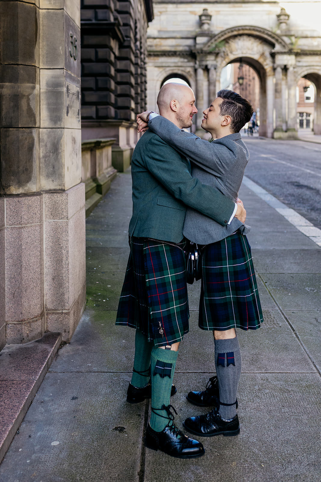 two grooms are facing each other and hugging each other. at the side of them there is a stone building and in the background there are the John Street arches.