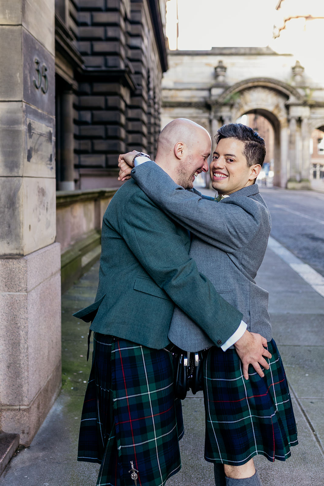 a couple are facing each other and hugging. one groom has his arms around his husbands neck. in the background you can see the john street arches.