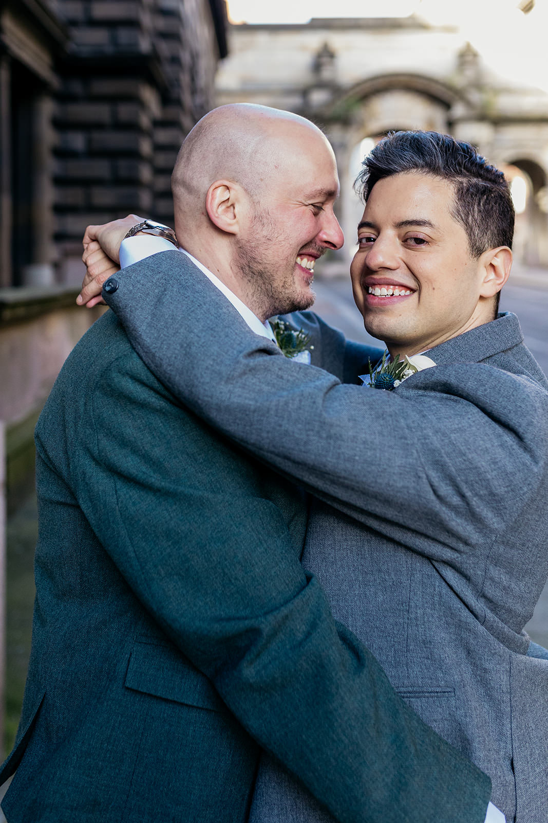 a couple are hugging each other and smiling. one groom is facing the camera