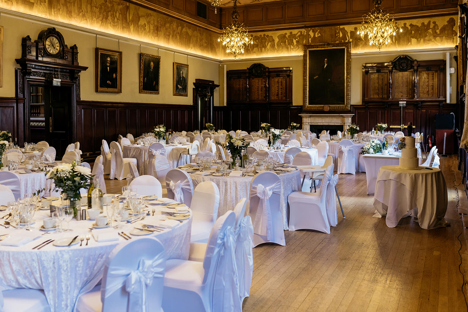 the trades hall is set up of the wedding breakfast. it is mostly white linens with white bows on the back of the chairs. chandeliers are hanging from the ceiling.