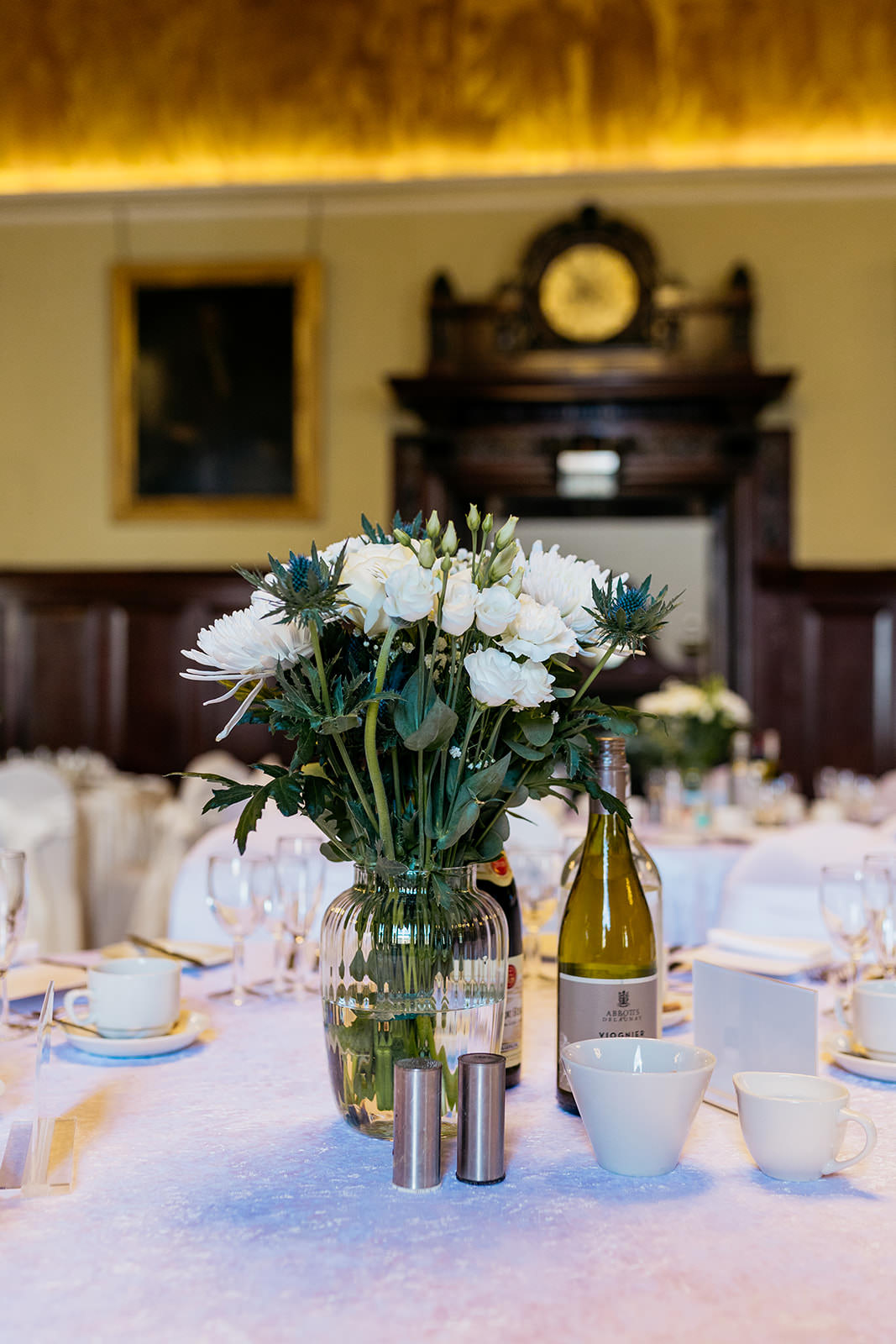 white flowers and blue thistles are gathered in a clear vase on the table to decorate for a wedding reception at Trades Hall. a bottle of white wine is next to the case.