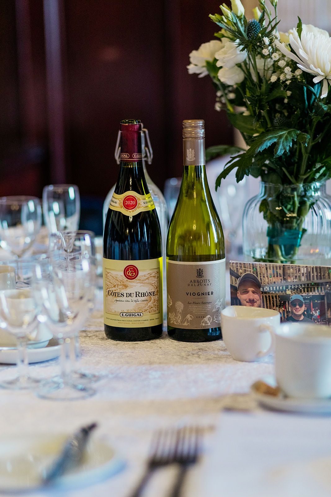 a bottle of red wine and white wine are sitting table at wedding reception. next to it is a vase of flowers and a picture of the two grooms.