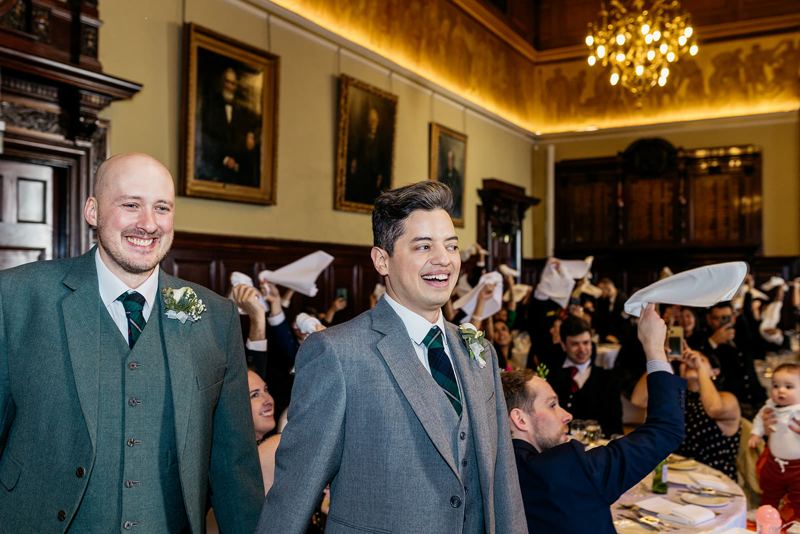 a happy couple are walking in to the trades hall the guests behind them are waving their napkins above their head.