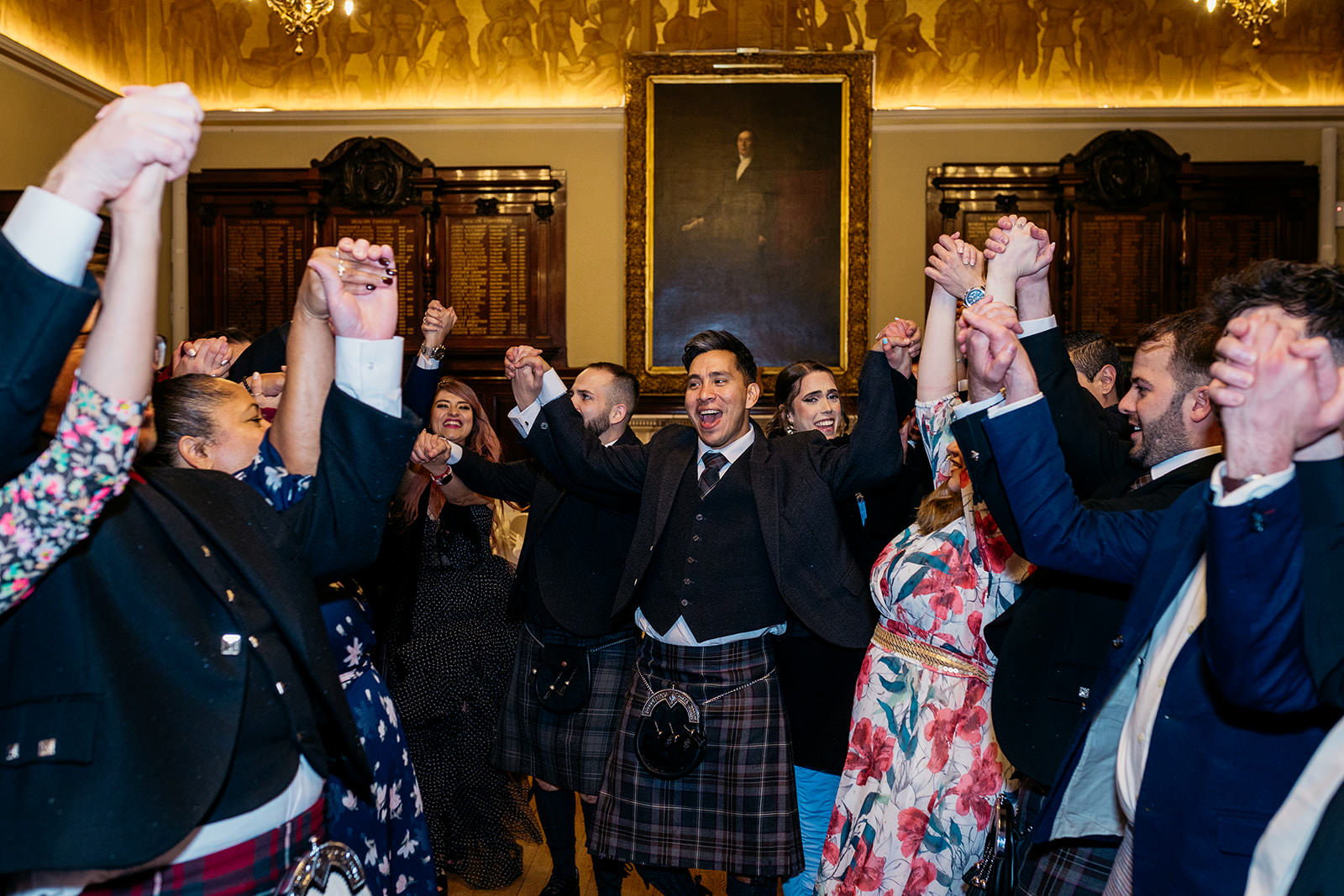 guests are ceilidh dancing at a wedding reception in Trades Hall, and they are all holding hands and cheering with their hands in the air.