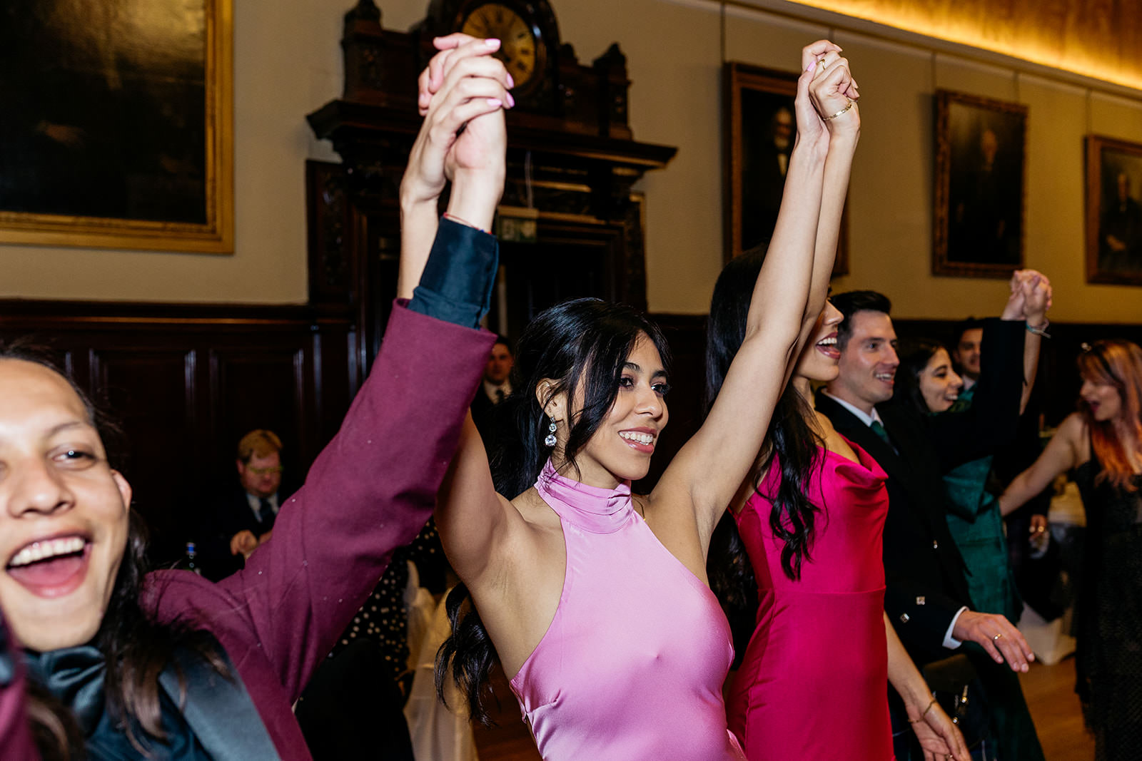 guests are ceilidh dancing at a wedding reception in Trades Hall, and they are all holding hands and cheering with their hands in the air.