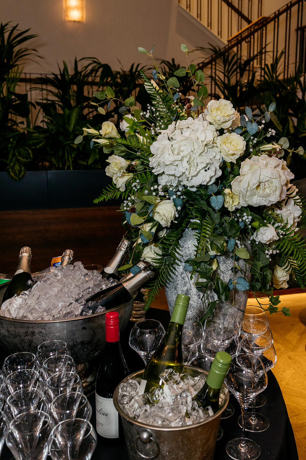 a large bouquet of flowers are in a glass vase on a table. There are buckets on the table filled with ice and champagne bottles.