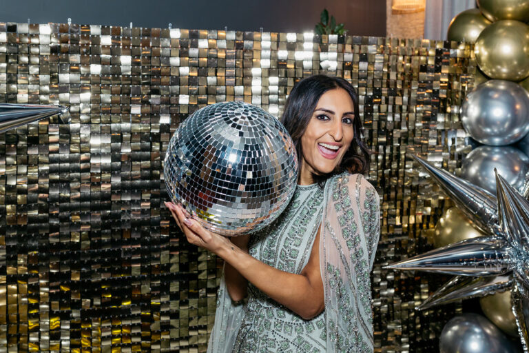 dr. punam krishan is holding a big glitter ball beside her face. she is standing in front of a gold glittery back drop