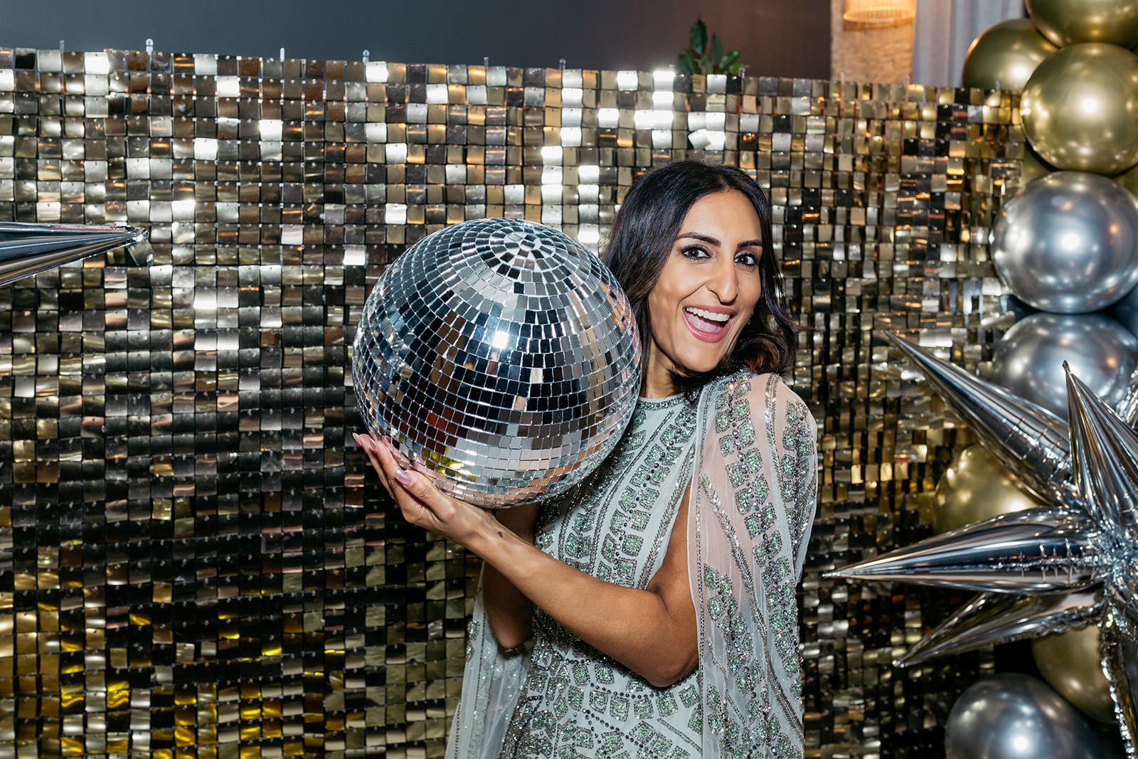 dr. punam krishan is holding a big glitter ball beside her face. she is standing in front of a gold glittery back drop
