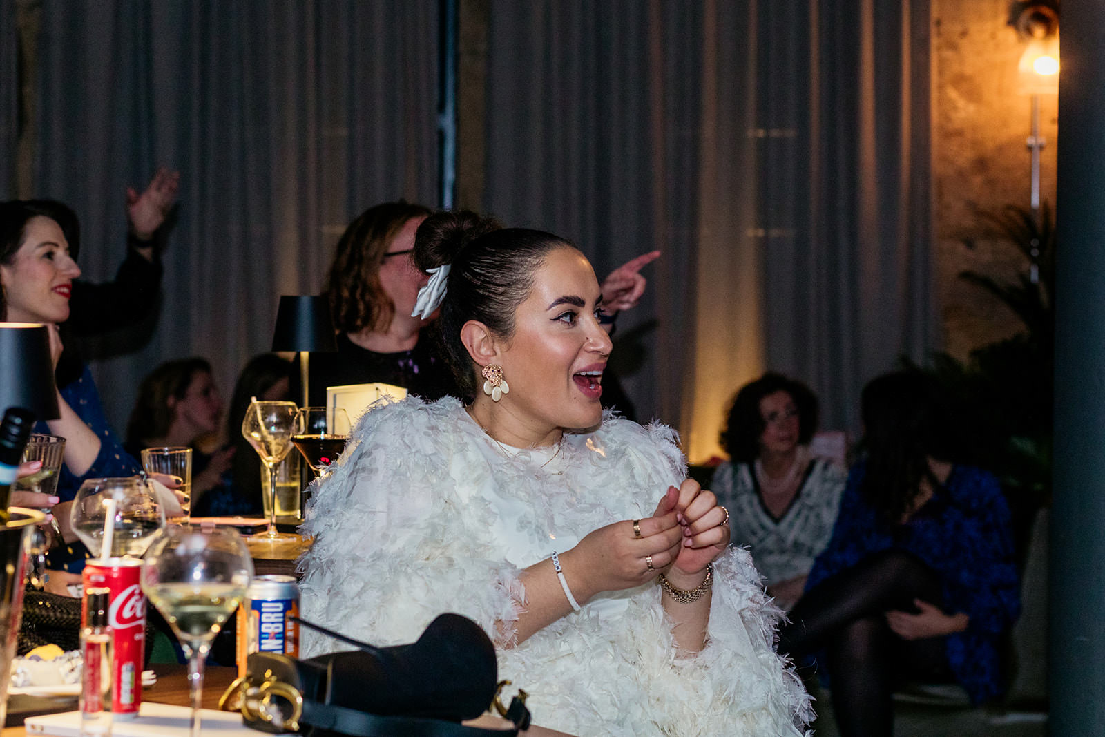 a woman wearing a white feathery top is smiling and in the background there are people pointing and smiling.