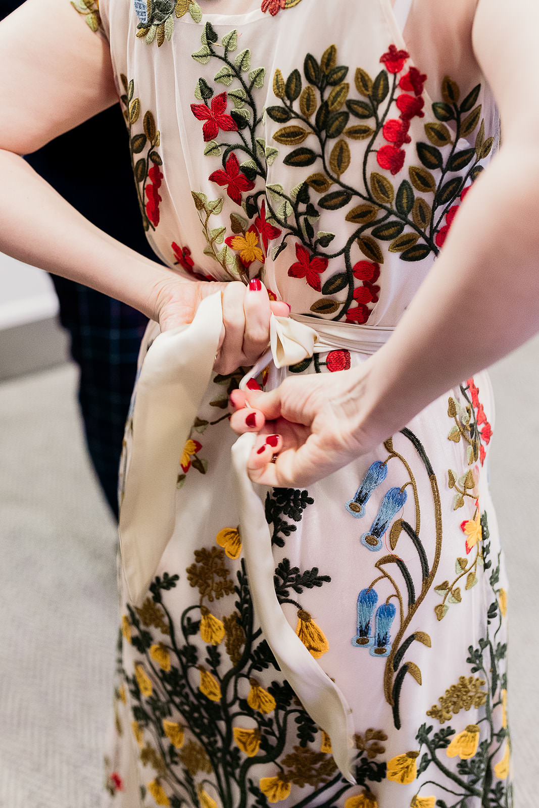 a woman is tying a belt behind her. the dress has a slip over the top of it that is covered with embroidered flowers.