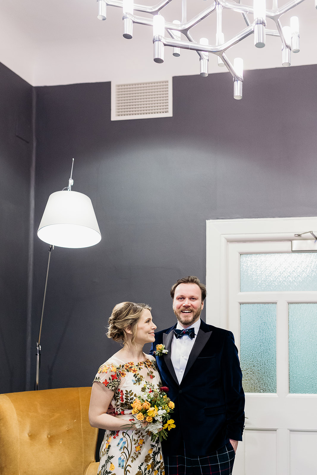 a bride is looking at the groom. they are standing in a room with dark grey walls, a mustard coloured chair and an overhanging lamp.