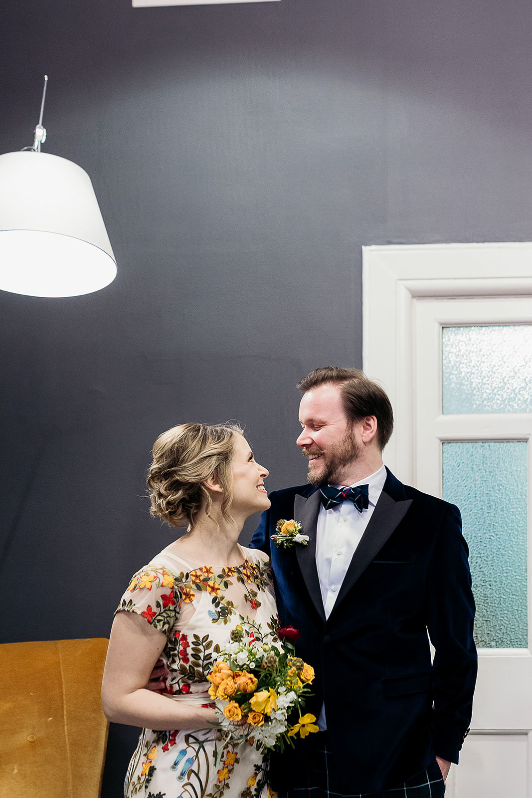 a bride and groom are looking at each other and smiling. they are in a room that has dark grey walls. they stand out because of the bright colours on the brides dress.