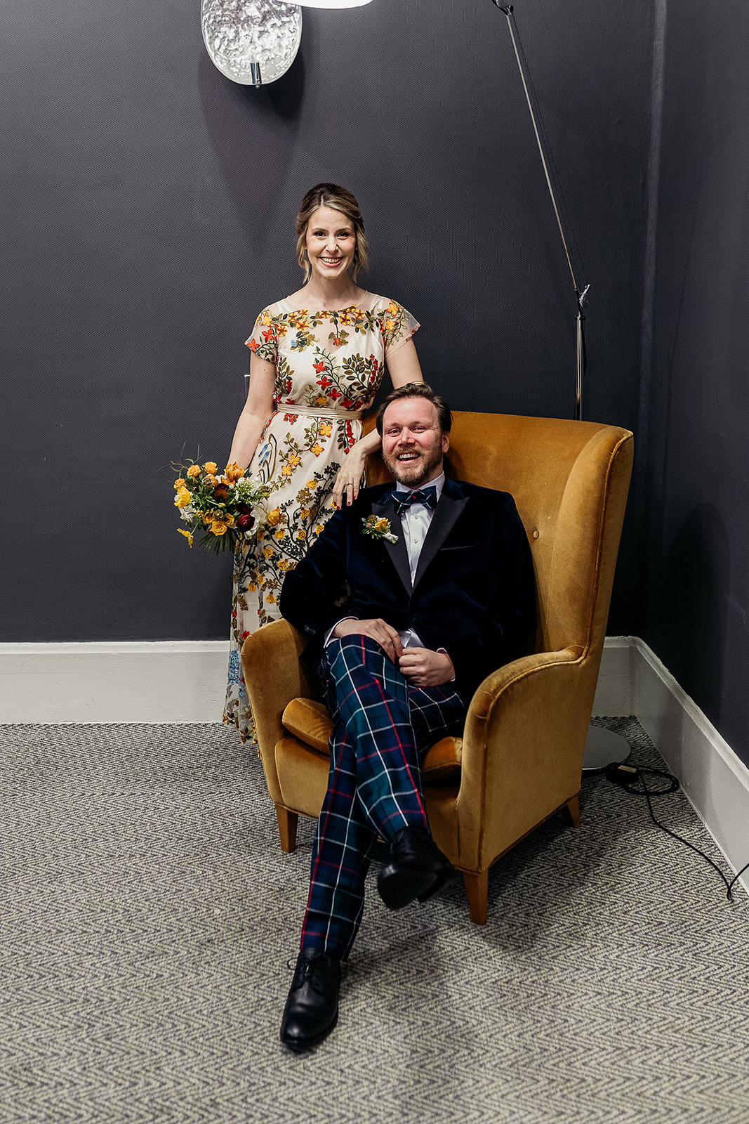 a man is sitting in a mustard coloured chair, and a woman is standing behind, leaning on the back. she is holding a colourful bouquet in her right hand.