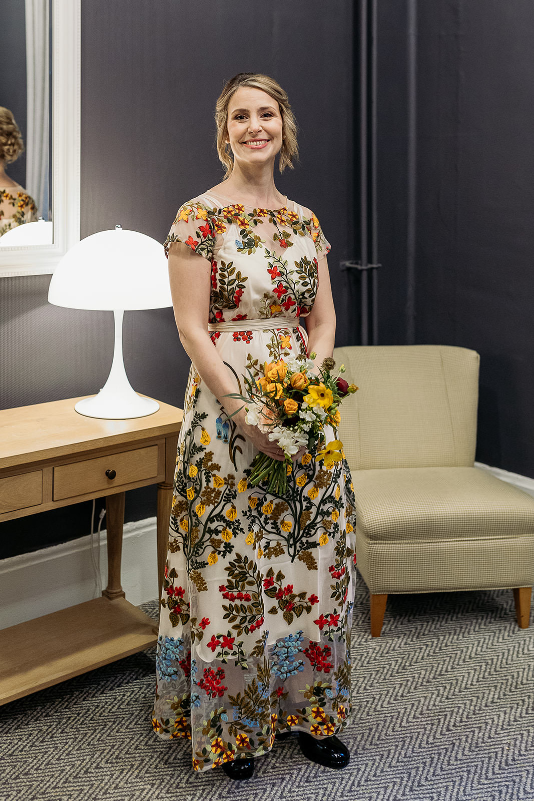 a woman is standing holding a bouquet. she is wearing a wedding dress that has layer covered with embroidered flowers. she is about to get married.