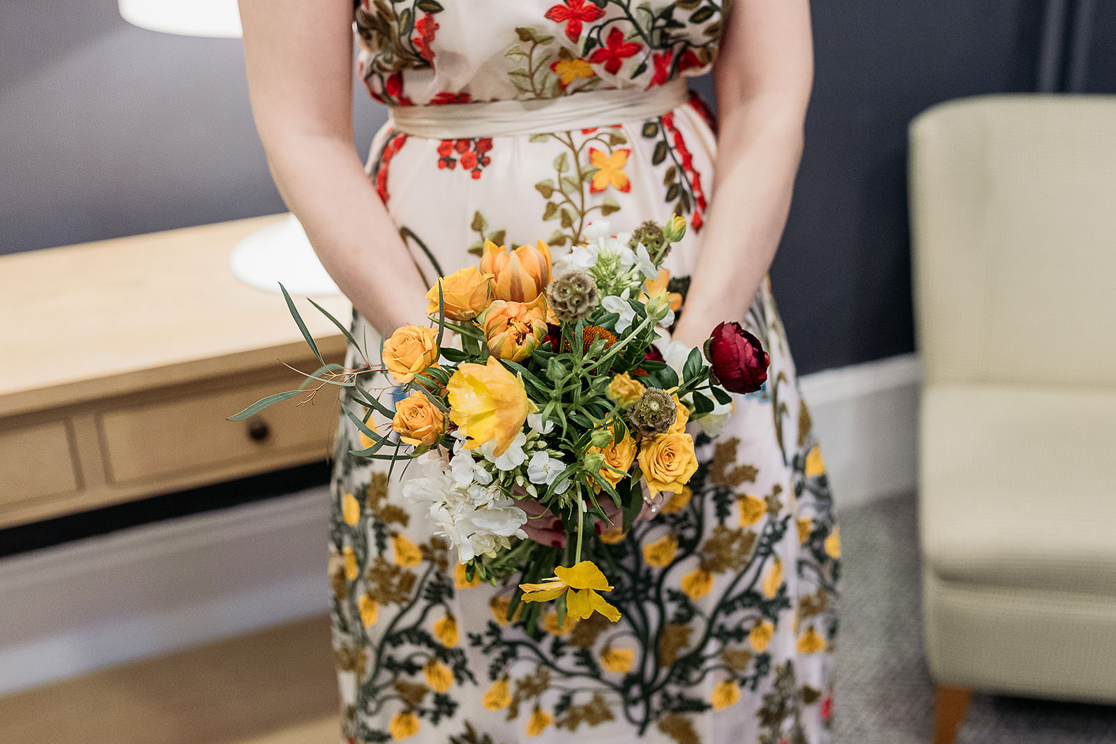 a colourful rustic bouquet which is filled with yellow flowers, green leaves, red and white. the woman is holding it in front of her embroidered dress.