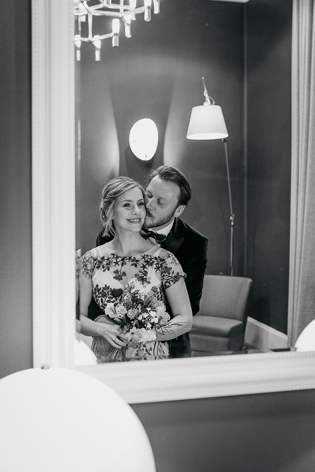 a man and woman are looking in to a mirror, he is standing behind her hugging in to her and kissing her cheek.