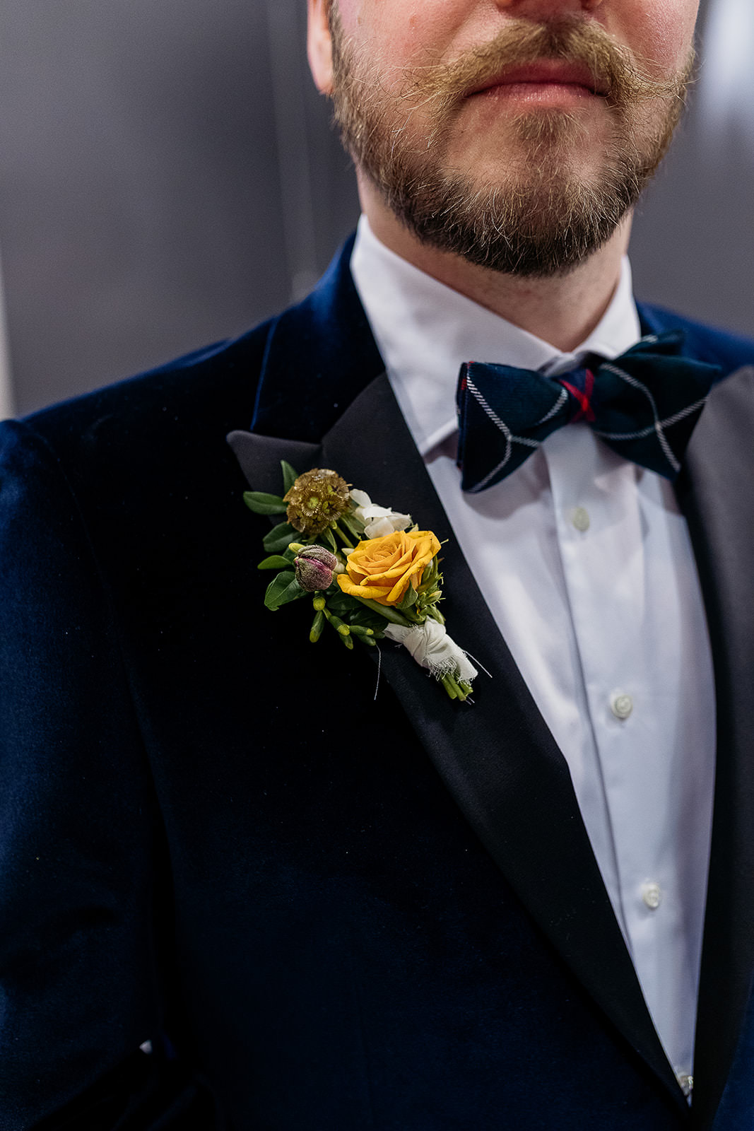 a boutonniere that is made up with a scabiosa flower, small yellow rose and small peony bud. it is placed on a dark blue velvet jacket, he is also wearing a white button wedding shirt and a tartan bow tie.